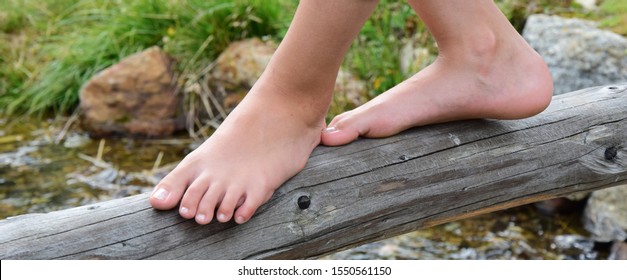 Young girl balances barefoot in the summer over a tree trunk that leads over a small stream
