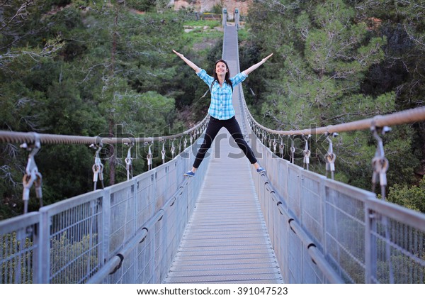 Young girl balancing on a railings of  suspension\
bridge through the forest in Haifa, Israel. challenge and win,\
Active life-stile concept. Young woman crossing the chasm on the\
rope pendant bridge