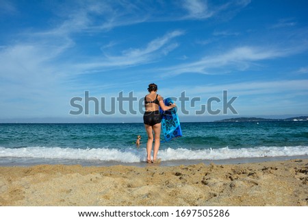 Young girl in black bikini on the beach, runs into the sea with a swimming board, a boy is in the water and the coast of the Cote d'Azur in the background