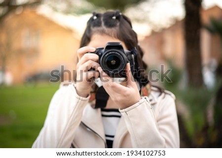 Young girl with black hair, Caucasian, dressed in beige windbreaker, black and white striped t-shirt, taking a picture with her reflex camera using a mask in a park