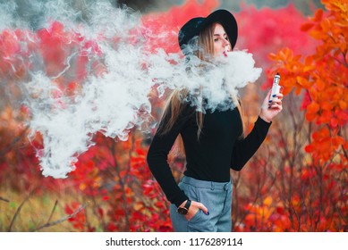 Young Girl In Black Hat Vaping Vape Device With Cloud Of Vape Outdoors In Autumn.