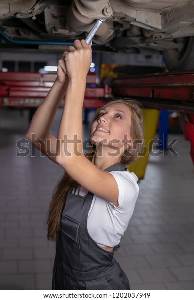 young girl in black jumpsuit and white\
t-shirt unscrews the nut with a wrench under the car and smiles.\
female mechanic works under the underside of a\
car