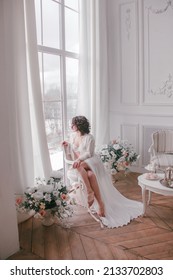 young girl with black short hair in white boudoir robe is sitting like bride in profile near huge panoramic window near white wall background with flowers. lifestyle concept, free space