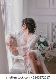 young girl with black short hair in white boudoir robe is sitting in profile near huge window on the white wall background with flowers. lifestyle concept, free space