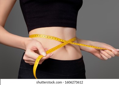 A young girl in black sportswear measures her waist with a centimeter on a dark gray background. Female body, slim waist. The girl’s healthy athletic body, diet, weight loss, calorie count. Slimming
