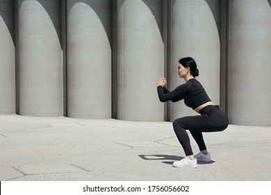 A young girl in a black tracksuit conducts street training and performs squats to strengthen the quadriceps
