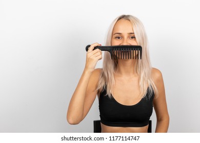 A young girl in a black T-shirt with blond hair shows a mustache on her face with a black comb. A photo on a white neutral homogeneous background in the studio.