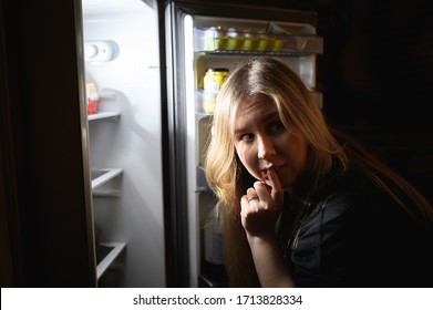 A Young Girl In A Black T-shirt With Long Blonde Hair, Stands Against The Open Refrigerator And Looks Back To Make Sure That No One Sees A Violation Of Her Diet. The Girl Is Alarmed