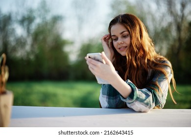 Young Girl Blogger Freelancer Holding Her Phone Looking At It And Working Outdoors In A Green Park Pensively Looking At The Screen Online