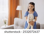 A young girl in a blue dress joyfully holds a plush toy while standing in a cozy living room setting. The ambiance is warm and inviting, highlighting feelings of innocence and contentment.