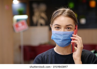 Young Girl In Blue Medical Protective Mask Talks On Smartphone Inside A Restaurant. Covid Concept Health And Safety, Coronavirus Quarantine, Social Distancing, Next Wave Covid Virus Protection