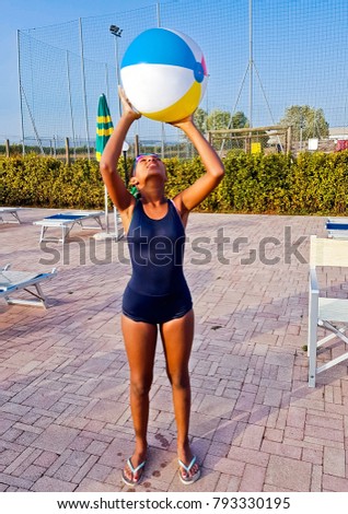 young girl in blue swimsuit plays with colorful ball in the swimming pool at sunset