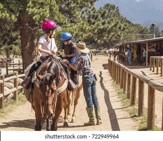Young girl and a boy on a horse listening to instructions of her horseback riding instructor; stables and woody mountains in the background; Colorado, USA