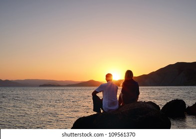 Young girl and boy sitting on a rock by the sea and watching the sunset