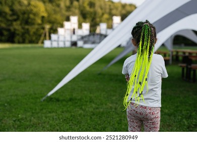 A young girl with bright green braided hair stands on a grassy field, wearing casual clothing, near a modern tent structure in a park setting on a sunny day. - Powered by Shutterstock