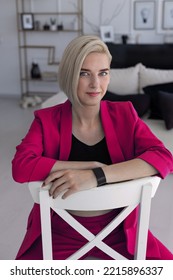 A Young Girl In A Bright Pink Suit Sits On A Chair In A Bright Room. Business Woman. Photo Session Of A Blonde Girl. Small Business Promotion.