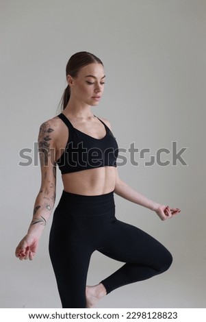 A young girl with brown hair, with a tattoo in a black top and leggings on a white background. Sport, fitness, standing with folded arms. Yoga