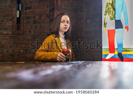 Young girl in a cafe, with a cup of coffee waiting for a meeting.In the background a brick wall and a stained glass colored window. Loft