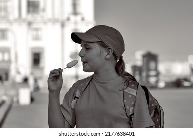 A Young Girl In A Cap And With A Backpack Behind Her Back Eats Ice Cream On The Street, Black And White Photo. A Child Cools Off With Popsicles Outdoors In The Summer.