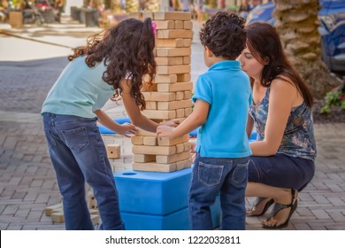 The Young Girl Is Careful Not To Knock Down The Wooden Large Size Jumbling Tower While Younger Brother Waits His Turn With Mommy. Mom Enjoys These Precious Moments Playing With Her Kids Outside.