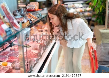 Young girl carefully examines the production in the department of the butcher shop in the supermarket, studying ..their cost