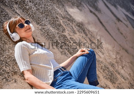 young girl in casual clothes and sunglasses lying on a sloping concrete surface listening to music with headphones on a sunny day
