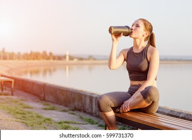 young girl of Caucasian appearance drinks a protein cocktail from a shaker after jogging in the open air. The bright sun illuminates it.