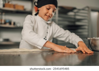 A young girl chef wearing a black headscarf and white chef coat is seen spreading dough on a stainless steel table in a professional kitchen environment with shelves in the background. - Powered by Shutterstock