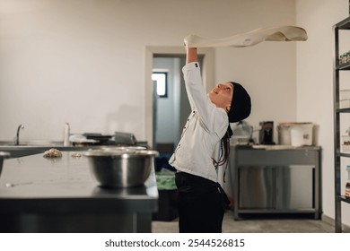 A young girl chef wearing a black headscarf and a white uniform practices tossing and stretching a sheet of dough in a professional kitchen, demonstrating artisanal bread-making techniques. - Powered by Shutterstock