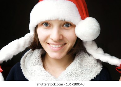 Young Girl Christmas Mood. The Girl Smiles In A New Year's Red Hat Pulled Over Her Eyes On A Dark Background.
