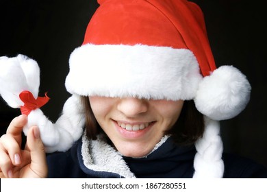 Young Girl Christmas Mood. The Girl Smiles In A New Year's Red Hat Pulled Over Her Eyes On A Dark Background.
