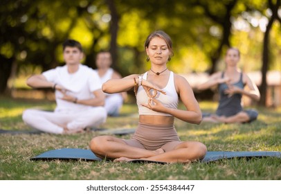 Young girl with closed eyes immersed in meditation during group yoga session in green sunlit summer park, sitting in lotus position, hands forming infinity sign with fingers folded in gyan mudra - Powered by Shutterstock