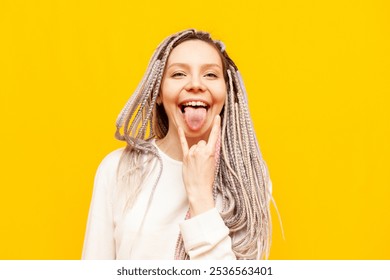 young girl with colored dreadlocks shows rock gesture and screams on yellow isolated background, crazy hipster woman with unique hairstyle and braids shows tongue