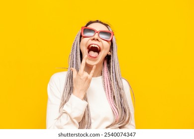 young girl with colored dreadlocks and sunglasses shows rock gesture and screams on yellow isolated background, crazy hipster woman with unique hairstyle and braids shows tongue