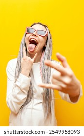 young girl with colored dreadlocks and sunglasses shows rock gesture and screams on yellow isolated background, crazy hipster woman with unique hairstyle and braids shows tongue