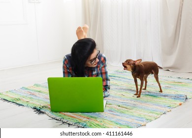 Young Girl In Computer Glasses Lying On Carpet With Laptop And Dog Chihuahua. Hipster Student, Freelancer Woman At Home. Relax With Computer. Girl With Barking Small Dog. Soft Tone, High Key.