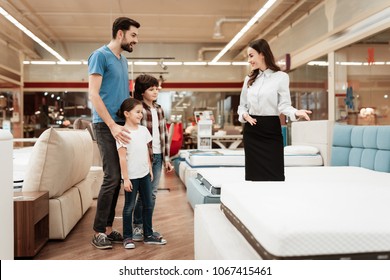 Young Girl Consultant Demonstrates Orthopedic Mattress To Young Father With Children In Furniture Store. Purchase Of Orthopedic Mattress. Sale Of Orthopedic Mattress In Furniture Store.