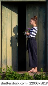 Young Girl In The Countryside In Summer. Violet Flower Is In Her Hands. In The Background Are Large Green Wooden Doors. Girl Stands Barefeet. Beautiful Young Woman Outdoors. Vacation In The Village.