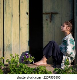 Young Girl In The Countryside In Summer. Violet Flower Is Near Her Legs. In The Background Are Large Green Wooden Doors. Girl Sits Barefeet. Beautiful Young Woman Outdoors. Vacation In The Village.