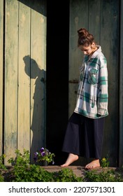 Young Girl In The Countryside In Summer. Violet Flower Is Near Her Legs. In The Background Are Large Green Wooden Doors. Girl Stands Barefeet. Beautiful Young Woman Outdoors. Vacation In The Village.