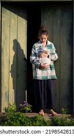 Young Girl In The Countryside In Summer. White-red Cat Is In Her Hands. In The Background Are Large Green Wooden Doors. Girl Stands Barefeet. Beautiful Young Woman Outdoors. Vacation In The Village.