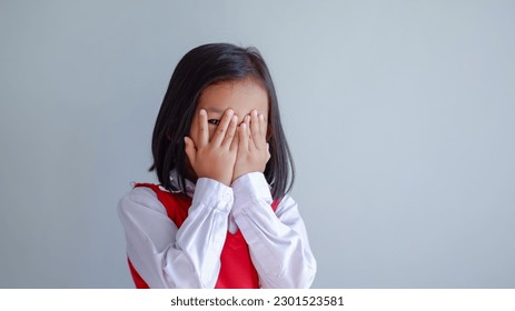 Young girl covering face with both hands being afraid of watching scary movie, peeping through hole between fingers. Confused timid child feeling shy hiding her emotions

