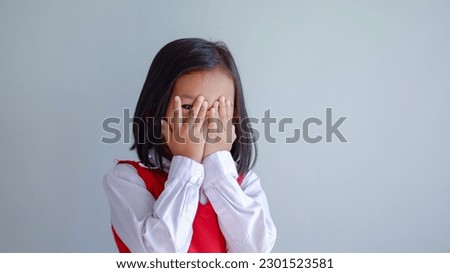 Young girl covering face with both hands being afraid of watching scary movie, peeping through hole between fingers. Confused timid child feeling shy hiding her emotions

