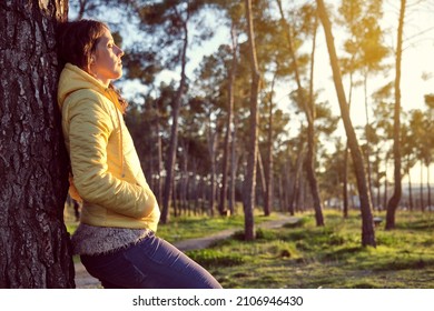 young girl with curly hair in yellow jacket and blue pants leaning on a pine tree with closed eyes enjoying nature in a pine forest at sunset