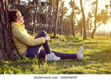 young girl with curly hair in yellow jacket and blue pants sitting on the ground leaning on a pine tree with her eyes closed enjoying nature in a pine forest at sunset