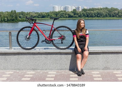 A Young Girl Cyclist Dressed In Bicycle Clothes Sit Down, Relax With A Red Road Bike With A View Of The River, Trees And Houses In The City.