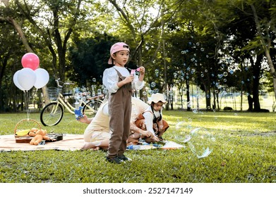 Young girl daughter blows bubbles during a family picnic in a sunny park. In the background, his family sits on a blanket, a picnic basket, and bicycles, enjoying a relaxing day outdoors. - Powered by Shutterstock