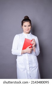 A young girl doctor in a white medical gown with a red book in her hands on a gray background - Shutterstock ID 2143480095