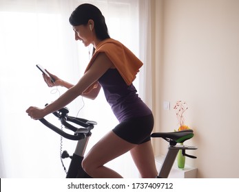 Young Girl Doing Exercise Bike At Home While Looking At The Smart Phone Profile View.