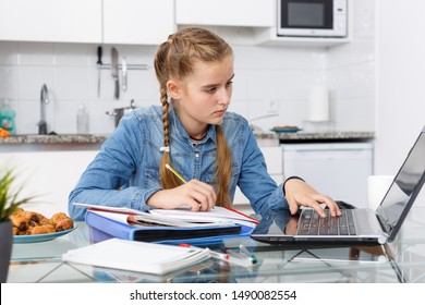 Сoncentrated young girl doing her homework with laptop and notebook at kitchen table